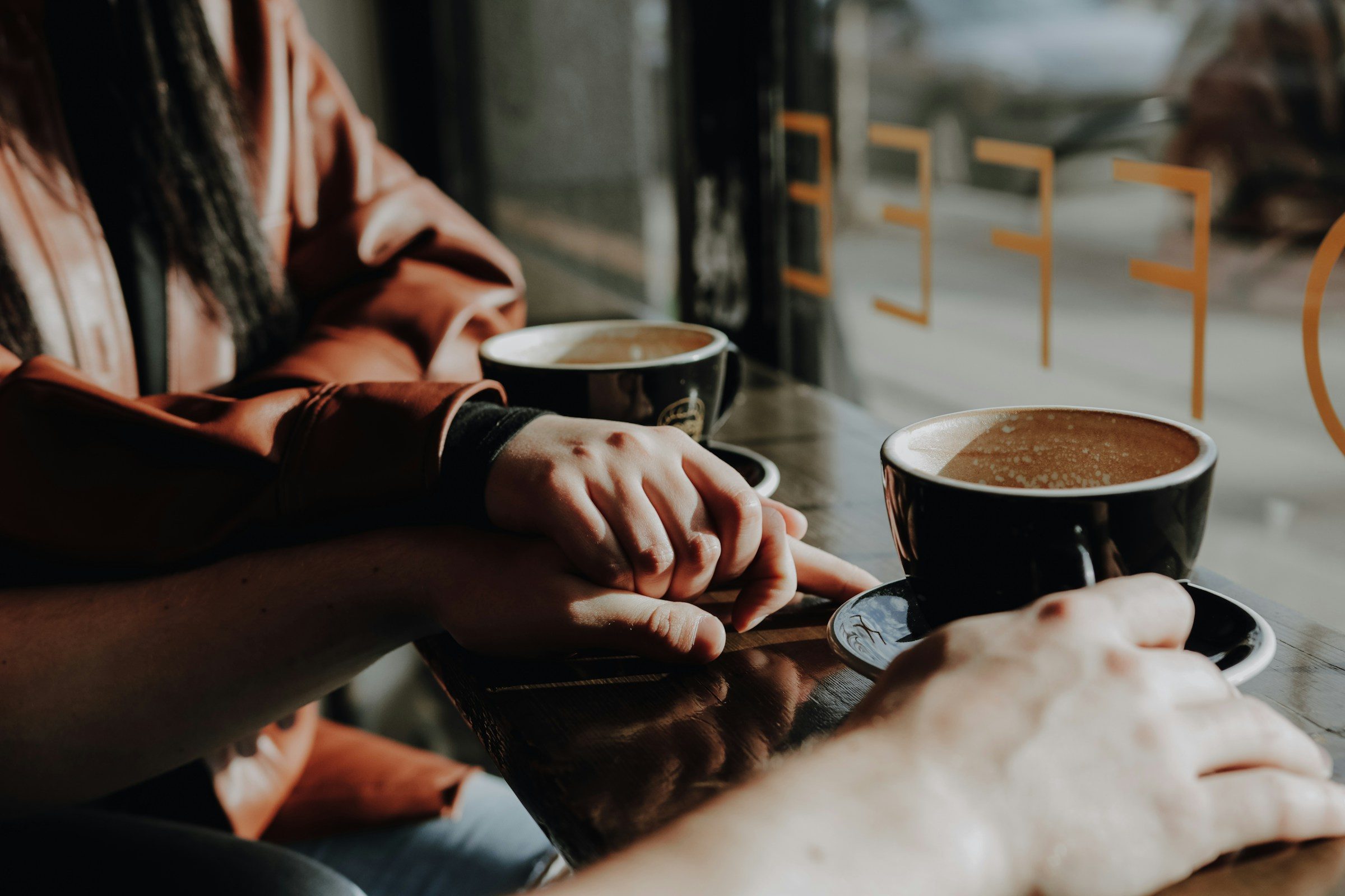Two people sitting across a cafe table with coffee, hands resting close together